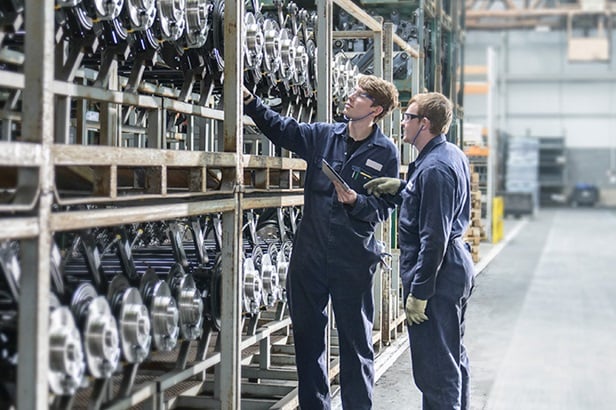 two men looking at rows of stored car axles on a shelving unit at a car plant